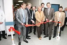 Eine Gruppe von Männern in Anzügen steht um einen roten Band. Sie bereiten sich darauf vor, das Band durchzuschneiden, um eine Eröffnung zu feiern. Im Hintergrund ist ein Krankenhausflur zu sehen.