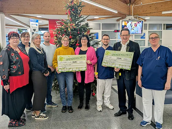 Gruppenfoto in einer Krankenhauslobby: Mehrere Personen, darunter Pflegekräfte in Kitteln, stehen vor einem geschmückten Weihnachtsbaum. Zwei Personen halten übergroße Spendenchecks hoch. Die Szene vermittelt eine Spendenübergabe zur Weihnachtszeit.