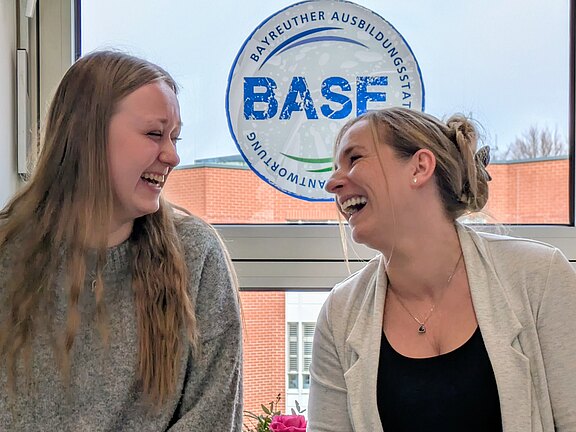 Zwei junge Frauen sitzen nebeneinander in einem Raum und lachen sich herzlich an. Tageslicht fällt durch ein Fenster. Die Szene vermittelt eine lockere, freundliche Stimmung wie bei einer entspannten Unterhaltung.