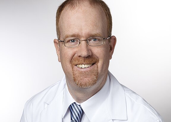 Porträt eines lächelnden Mannes mit Brille und rotem Bart, der einen weißen Kittel und eine gestreifte Krawatte trägt, vor hellem Hintergrund.