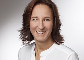 Porträtfoto: Eine lächelnde Frau mit schulterlangem braunem Haar blickt in die Kamera. Sie trägt eine weiße Bluse vor neutralem, hellgrauem Hintergrund; freundlicher, professioneller Ausdruck.