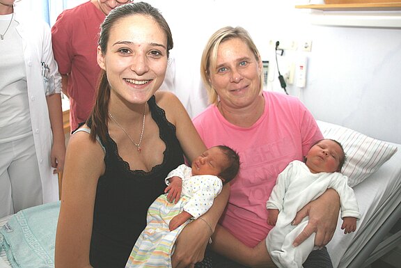Zwei Frauen sitzen in einem Krankenzimmer und lächeln. Jede hält ein neugeborenes Baby im Arm, als ob die Geburt gerade stattgefunden hätte.