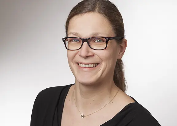 Porträt einer lächelnden Frau mit dunkler Brille, Haare zum Zopf gebunden, schlichtes schwarzes Oberteil vor neutral hellem Hintergrund.