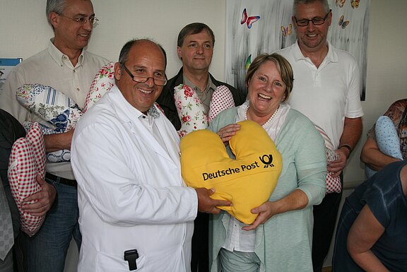 Mehrere lächelnde Erwachsene; ein Arzt im weißen Kittel und eine Frau halten gemeinsam ein gelbes, herzförmiges Kissen mit der Aufschrift „Deutsche Post“. Weitere Personen im Hintergrund tragen ähnliche Herzkissen.