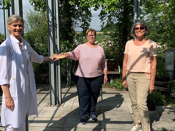 Drei Frauen stehen im Freien. Links trägt eine Frau einen weißen Kittel und überreicht der mittleren Frau einen Umschlag. Die dritte Frau steht daneben und lächelt.