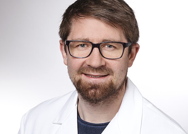 Porträt eines lächelnden Mannes mit Brille, braunem Kurzhaar und Bart. Er trägt einen weißen Laborkittel über dunklem Shirt. Heller, neutraler Hintergrund.