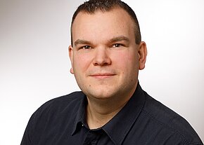 Johannes Sperber Porträtfoto einer Person mit kurzem dunklem Haar und leichtem Lächeln, die direkt in die Kamera blickt. Sie trägt ein dunkelgraues Hemd; der Hintergrund ist hell und neutral, wie bei einem professionellen Bewerbungsfoto.