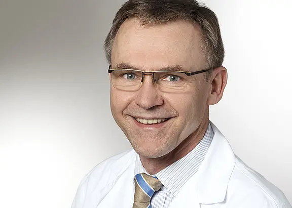 Porträt eines lächelnden Mannes mittleren Alters mit Brille im weißen Arztkittel, Hemd und gestreifter Krawatte vor neutralem Hintergrund.