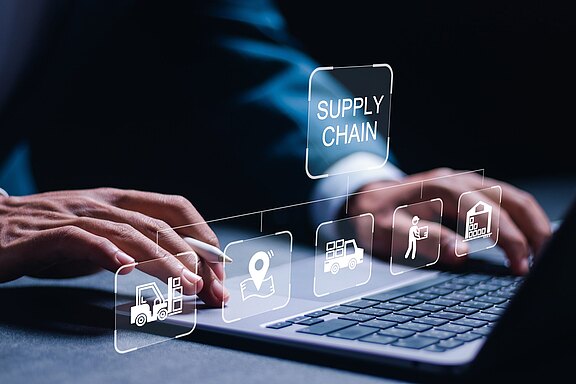 Hände tippen auf einem Laptop; darüber schwebt der Begriff „SUPPLY CHAIN“ mit Symbolen für Logistikprozesse: Gabelstapler, Standortmarke, Liefer-Lkw, Mitarbeiter mit Paket und Lagerhaus. Visualisierung digitaler Lieferkettensteuerung.