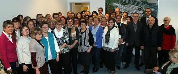 Große Gruppe von Erwachsenen posiert in einem Innenraum für ein Gruppenfoto. Die meisten lächeln; viele Frauen, einige Männer, gemischte Altersgruppen. Locker bis geschäftlich gekleidet, stehen dicht beieinander vor heller Wand. Stimmung freundlich, Anlass wirkt wie Treffen oder Feier.
