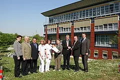 Eine Gruppe von acht Personen steht vor einem modernen Gebäude. Einige tragen Arbeitskleidung, darunter eine Krankenschwester. Sie scheinen an einer Übergabe oder Präsentation beteiligt zu sein, da sich einige Hände zur Begrüßung oder zum Austausch reichen.