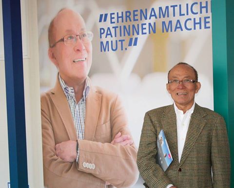 Ein lächelnder Mann steht neben einem großen Plakat, auf dem er ebenfalls abgebildet ist. Der Hintergrund des Plakats ist schlicht und trägt den Text „Ehrenamtlich Patinnen machen Mut.“ Der Mann trägt eine Brille und hält eine Broschüre in der Hand.