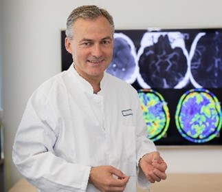 Ein lächelnder Arzt in einem weißen Kittel steht vor einem Bildschirm, auf dem verschiedene medizinische Bildgebungen des Gehirns zu sehen sind. Er sieht aus, als ob er gerade eine Erklärung oder Diskussion vorbereitet.