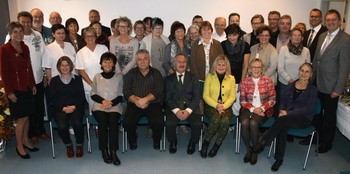 Gruppenfoto in einem Innenraum: Etwa dreißig Erwachsene posieren für eine formelle Aufnahme, erste Reihe sitzt, dahinter mehrere Reihen stehen. Alle blicken zur Kamera und lächeln. Kleidung wirkt beruflich bis festlich. Die Szene vermittelt den Eindruck eines Team- oder Jubiläumstreffens.