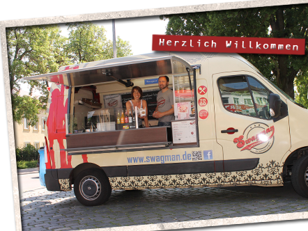 Ein Food-Truck mit der Aufschrift "Swagman" bietet verschiedene Getränke an. Zwei Personen stehen mit einem freundlichen Lächeln hinter der Theke. Über dem Truck befindet sich ein Schild mit der Begrüßung "Herzlich Willkommen". Der Truck steht auf einem gepflasterten Platz, umgeben von Bäumen.