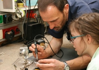 Ein Erwachsener zeigt einem Mädchen am Werktisch, wie man elektronische Bauteile lötet. Beide beugen sich konzentriert über eine kleine Schaltung; der Mann führt den Lötkolben, das Kind beobachtet aufmerksam. Lern- und Reparatursituation in einer Werkstatt.