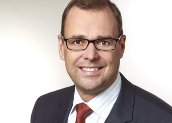 Porträt eines lächelnden Mannes mit Brille, im dunklen Anzug und gestreiftem Hemd mit roter Krawatte, vor hellem, neutralem Hintergrund.