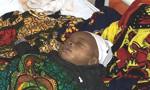 Ein schlafendes Baby liegt auf dem Rücken, in bunte gemusterte Stoffe gewickelt. Es trägt eine weiße Mütze, das Gesicht wirkt ruhig und entspannt.