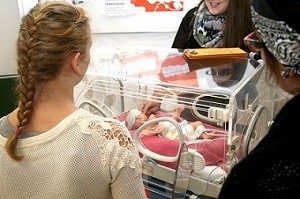 Eine Frau mit geflochtenem Haar steht vor einem Inkubator, in dem ein Neugeborenes liegt. Im Hintergrund sprechen zwei weitere Frauen miteinander. Die Szene vermittelt den Moment der Beobachtung und des Austausches über das Baby im Inkubator.