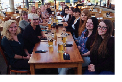 Eine Gruppe von zwanzig Personen sitzt an einem langen Tisch in einem Restaurant. Vor ihnen stehen Gläser mit Bier. Die Atmosphäre wirkt gesellig und einladend. Die Frauen und Männer lächeln und scheinen an einem gemeinsamen Ereignis oder Treffen teilzunehmen.