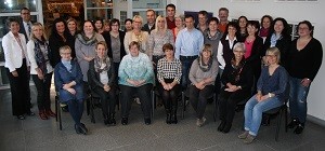 Großes Gruppenfoto: Rund 30 Erwachsene posieren in einem Innenraum. Erste Reihe sitzt auf Stühlen, hintere Reihe steht. Alle blicken zur Kamera, freundliche Stimmung, Teamfoto.