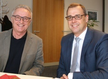 Zwei Männer sitzen lächelnd an einem Tisch in einem Büro. Der linke trägt ein graues Sakko, der rechte einen blauen Anzug mit Krawatte. Beide tragen Brillen.