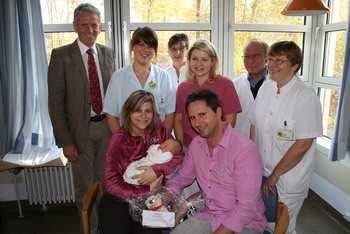 Eine stolze Familie hält ein Neugeborenes in den Armen, umgeben von einer Gruppe von medizinischem Personal. Die Szene vermittelt Freude und Unterstützung in einem Krankenhausumfeld.