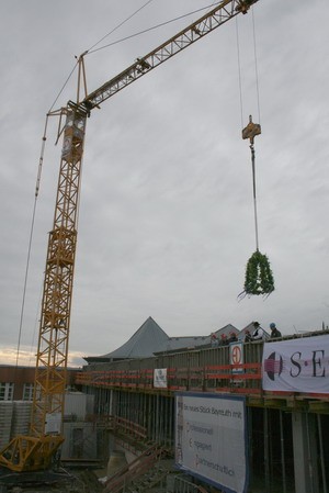 Ein Kran hebt einen Kranz über eine Baustelle, während Arbeiter auf dem Gerüst stehen. Im Hintergrund sind Teile eines Gebäudes und Werbebanner zu sehen. Der Himmel ist bewölkt und sorgt für eine düstere Atmosphäre.
