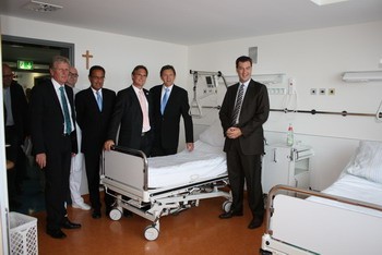 Sechs Männer stehen in einem Krankenhauszimmer mit zwei leeren Betten. Sie tragen formelle Kleidung und lächeln. Im Hintergrund sind medizinische Geräte und helle Wände zu sehen, was eine moderne Krankenhausumgebung vermittelt.