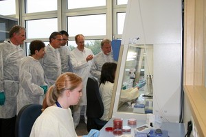 In einem Labor arbeitet eine Frau am Tisch, während mehrere Personen in weißen Laborkitteln von ihr lernen. Die Gruppe schaut aufmerksam zu, während sie an einem Experiment beteiligt ist.