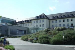 Ein mehrstöckiges Gebäude mit moderner Glasarchitektur ist im Vordergrund zu sehen. Im Hintergrund ist ein traditionellerer Gebäudeteil mit einem Satteldach. Um das Gebäude herum gibt es eine grüne Landschaft mit Blumen und Büschen.