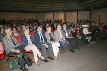 Eine Gruppe von Menschen sitzt auf roten Stühlen in einem Saal. Die Zuschauer schauen aufmerksam in Richtung Bühne, wobei einige Personen im Vordergrund gut zu erkennen sind. Der Raum ist voll mit einer großen Anzahl von Teilnehmern, die an einer Veranstaltung oder Präsentation teilnehmen.