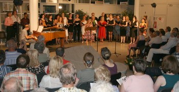 Eine Chorgemeinschaft steht auf einer Bühne und singt, während ein Pianist am Klavier begleitet. Das Publikum, bestehend aus älteren Menschen, sitzt in Reihen und hört aufmerksam zu. Die Atmosphäre ist festlich und musikalisch.