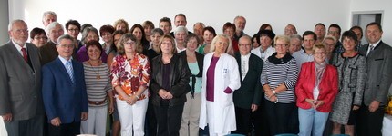 Das Bild zeigt eine große Gruppe von Menschen, die zusammenstehen und in die Kamera schauen. Es sind etwa 50 Personen, darunter Frauen und Männer verschiedenen Alters, einige in Arbeitskleidung, darunter auch eine Person in einem weißen Kittel. Die Szene wirkt freundlich und einladend.