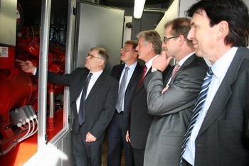 Eine Gruppe von fünf Männern in Anzügen steht vor einem technischen Gerät. Ein Mann zeigt auf die Maschine, während die anderen aufmerksam beobachten. Die Szene vermittelt Interesse und Diskussion über die Technologie im Hintergrund.