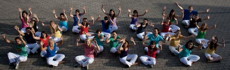 Eine Gruppe von etwa 30 Menschen sitzt auf dem Boden, trägt bunte Oberteile und weiße Hosen. Sie lächeln, heben die Arme und wirken fröhlich. Die Pose vermittelt ein Gefühl von Gemeinschaft und Freude.