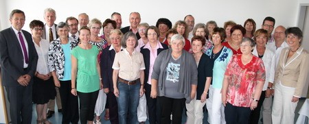 Eine Gruppe von etwa 40 Menschen steht in einem Raum. Die Mehrheit sind ältere Frauen, die in verschiedenen Farben gekleidet sind. Einige Männer sind ebenfalls anwesend. Alle schauen in Richtung der Kamera und lächeln. Der Hintergrund ist hell und neutral.