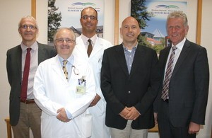 Fünf männliche Personen stehen zusammen in einem Büro, wobei zwei in medizinischen weißen Kitteln gekleidet sind. Im Hintergrund sind Plakate sichtbar, die medizinische Einrichtungen präsentieren. Die Gruppe scheint freundlich und professionell.