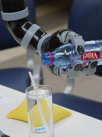 Ein robotischer Arm greift eine Plastikflasche mit Wasser. Die Flasche steht dabei über einem Glas Wasser mit einem Strohhalm. Der Arm zeigt eine flexible Hand mit Greifmechanismus, die darauf ausgelegt ist, Objekte zu halten und zu manipulieren.