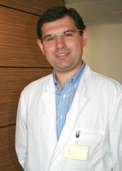 Ein Mann in einem weißen Kittel steht vor einer Wand und lächelt. Er trägt eine Brille und ein blau gestreiftes Hemd darunter. An seinem Kittel befestigt ist ein Namensschild. Die Umgebung wirkt professionell und freundlich.