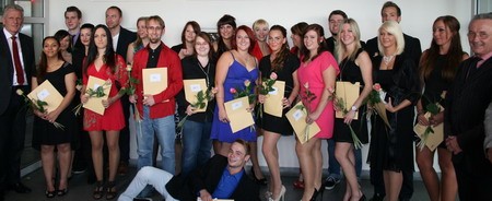 Eine Gruppe von Personen steht zusammen und hält Urkunden sowie Rosen in den Händen. Sie scheinen an einer Feier teilzunehmen, möglicherweise einer Abschlusszeremonie. Einige Teilnehmer lächeln und posieren freundlich für das Foto.