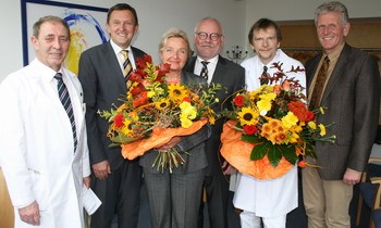 Sechs Personen stehen zusammen in einem Raum, halten große Blumensträuße mit bunten Blumen. Die Männer tragen Anzüge und der Arzt einen weißen Kittel. Die Stimmung ist festlich, da es sich um eine besondere Gelegenheit handelt.
