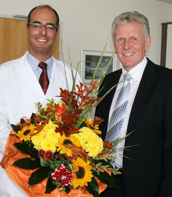 Zwei Männer stehen nebeneinander und lächeln. Der Mann links trägt einen weißen Kittel und hält einen großen Blumenstrauß mit bunten Blumen in den Händen. Der Mann rechts trägt einen Anzug und sieht erfreut aus. Beide haben eine positive Ausstrahlung.