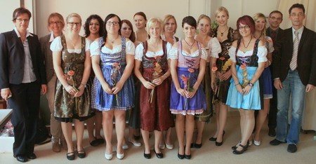 Eine Gruppe von 14 Frauen und 2 Männern steht zusammen. Die Frauen tragen traditionell bayerische Dirndl in verschiedenen Farben, häufig mit Blumen. Einige halten eine Blume in der Hand. Der Hintergrund ist neutral, und alle Personen lächeln freundlich in die Kamera.
