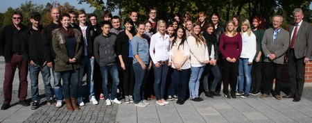 Eine Gruppe von etwa 30 Personen steht im Freien auf einem Platz. Sie sind gemischt in Geschlecht und Alter und tragen unterschiedliche Kleidung. Im Hintergrund sind Bäume und der Himmel sichtbar. Einige Personen lächeln zur Kamera.