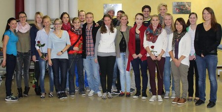 Eine Gruppe von etwa 20 jungen Erwachsenen steht zusammen in einem Raum. Einige halten Blumen. Die Personen lächeln und sind in unterschiedlichen, lässigen Kleidungsstilen gekleidet. Im Hintergrund sind dekorative Elemente sichtbar.