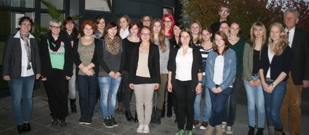 Eine Gruppe von 20 Personen steht in einem Innenhof. Die Mehrheit sind Frauen unterschiedlichen Alters, die lächeln und in casual Kleidung gekleidet sind. Im Hintergrund sind Pflanzen und ein Gebäude sichtbar. Ein Mann steht am rechten Rand der Gruppe.