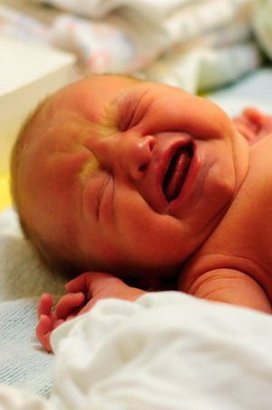 Ein weinendes Neugeborenes liegt auf einer weichen Unterlage. Das Baby hat eine gerötete Haut und scheint unruhig zu sein, während es seine Hände in einer geballten Faust hält. Das Bild vermittelt den emotionalen Ausdruck des Babys in diesem Moment.