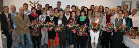 Eine Gruppe von etwa 30 Personen steht zusammen, die meisten halten Blumensträuße. Sie lächeln und posieren vor einer hellen Wand. Im Hintergrund ist ein Weihnachtsbaum zu sehen. Die Atmosphäre wirkt feierlich und herzlich.