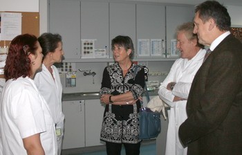 Fünf Personen, darunter Ärzte in weißen Kitteln, stehen in einem Krankenhausraum. Eine Frau in einem schwarzen Oberteil spricht mit den anderen. Alle scheinen sich angeregt zu unterhalten, während sie um eine Arbeitsfläche herum stehen.